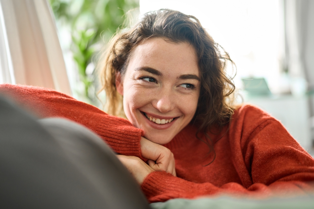 Happy relaxed calm beautiful lady enjoying comfort thinking or good on soft sofa looking away. Young smiling pretty curly woman relaxing sitting on couch at home and dreaming. Close up.